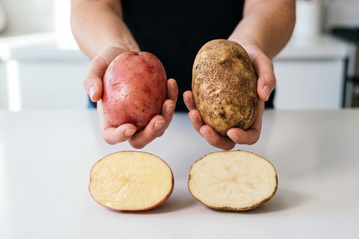 Cut halves of a red waxy potato and a brown starchy potato placed side by side on a white surface.