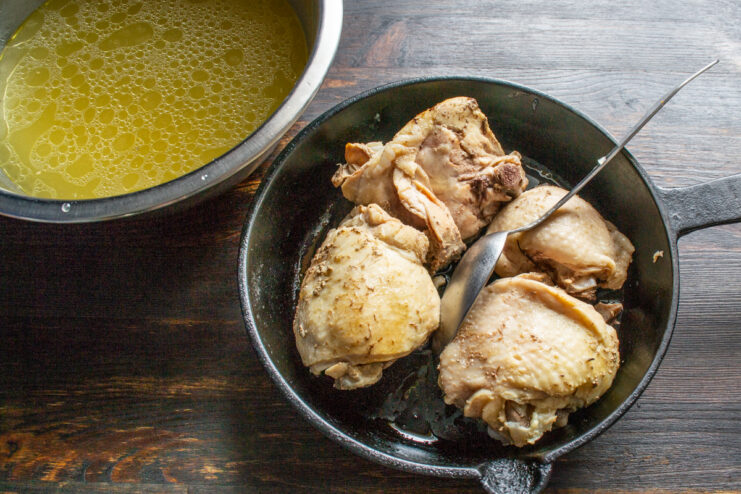 Prepping chicken for the final step Boiled chicken pieces in a pan next to a bowl of clear broth