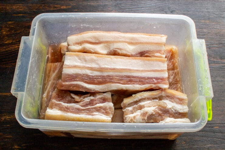 Layering raw pork belly pieces with salt and sugar in a curing container
